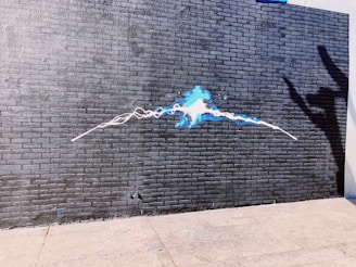 A brick wall painted dark gray with a vibrant blue and white spray paint art in the center. The artwork resembles an electric pulse or abstract energy flowing horizontally across the wall. To the right, a shadow of a hand creates a playful or dramatic effect.