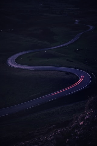 A winding road meanders through a dark, mountainous landscape, with faint light trails indicating the movement of a vehicle. The surrounding scenery is shadowy and subdued, creating a sense of isolation and tranquility.