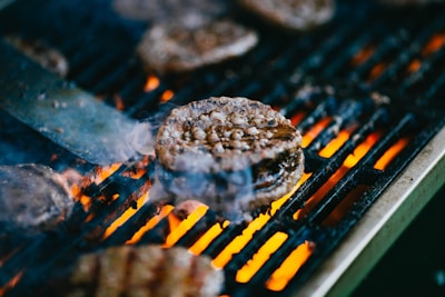 Juicy burger patties cooking on a flaming grill, with visible spatula and rising smoke adding to the sense of a freshly grilled meal.