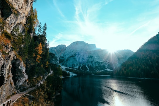 A serene mountain landscape with a lake reflecting the sunlight and a trail running along the rocky cliffs on the left. The trees are a mix of green and autumnal shades, and the sun is peeking over the mountains in the background, creating a dramatic sunburst effect.