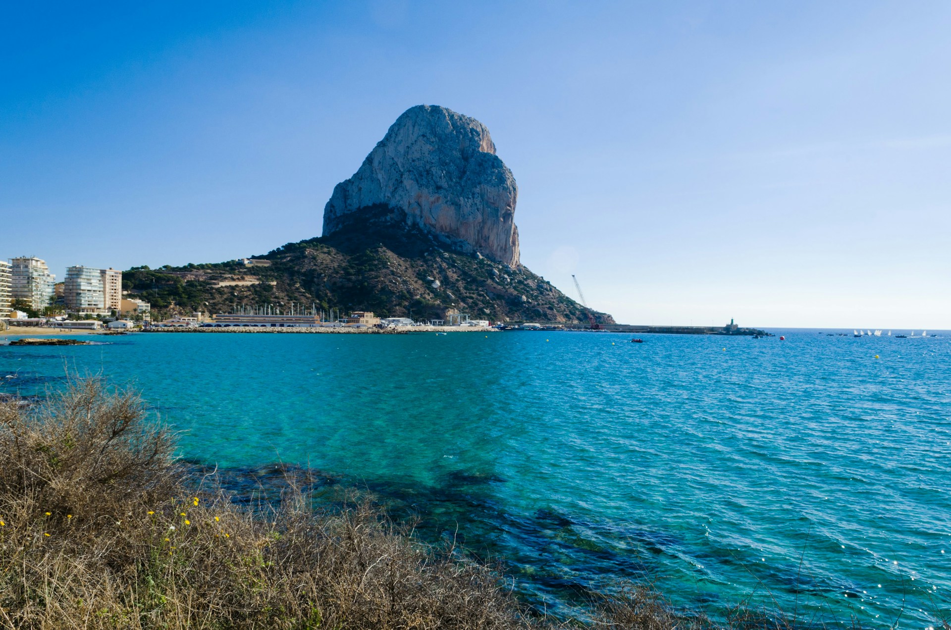A large rocky mountain dominates the background, rising dramatically from a coastal town with several modern buildings. The clear blue sea extends towards the foreground, with small boats visible near the horizon. Sparse vegetation and a few yellow flowers are in the foreground, contrasting with the blue water.