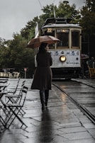 A person holding an umbrella walks on a wet cobblestone street towards an approaching vintage tram. The tram is labeled as route 22 to Batalha and features decorative designs on its exterior. Empty outdoor café tables and chairs are visible on the side, and there are lush green trees in the background. The atmosphere is overcast and rainy.