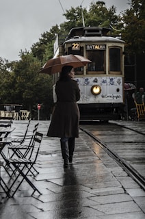 A person holding an umbrella walks on a wet cobblestone street towards an approaching vintage tram. The tram is labeled as route 22 to Batalha and features decorative designs on its exterior. Empty outdoor café tables and chairs are visible on the side, and there are lush green trees in the background. The atmosphere is overcast and rainy.