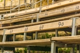 Wooden bleacher seats are arranged in rows with numbers painted on them. The setting appears outdoors, as indicated by greenery visible below the structure. Metal supports are used to hold the wooden planks in place, providing a rustic and aged appearance.