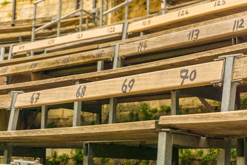 Wooden bleacher seats are arranged in rows with numbers painted on them. The setting appears outdoors, as indicated by greenery visible below the structure. Metal supports are used to hold the wooden planks in place, providing a rustic and aged appearance.