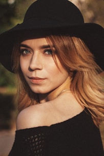 A young woman with light brown hair wears a wide-brimmed black hat and black off-shoulder top. She gazes directly at the camera with a soft expression. The background is softly blurred, highlighting her features with natural lighting that casts gentle shadows on her face.