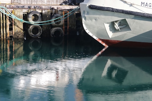 A docked boat with a visible bow and part of a pier in the background. The water reflects the boat and pier, and there are rubber tires attached to the pier, likely serving as bumpers.