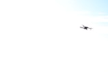 A drone is flying in the sky, with a slightly tilted angle. The background is predominantly white, possibly overexposed, with hints of light blue indicating a clear sky.