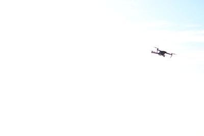 A drone is flying in the sky, with a slightly tilted angle. The background is predominantly white, possibly overexposed, with hints of light blue indicating a clear sky.