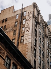 Tall, old brick buildings with many windows and a large vertical sign that reads 'Robert Beatson Collection'. The sign advertises handbags and accessories, and there are other architectural details with ornate decorations. The sky is partly cloudy.