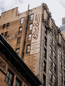 Tall, old brick buildings with many windows and a large vertical sign that reads 'Robert Beatson Collection'. The sign advertises handbags and accessories, and there are other architectural details with ornate decorations. The sky is partly cloudy.