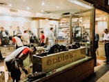 A busy shoeshine and repair store featuring multiple people getting their shoes shined by workers wearing red shirts. Shelves filled with shoes and repair equipment are visible, along with a storefront displaying 'Eddie’s Shoeshine & Repair'. The atmosphere is lively with a mix of customers seated and staff attending to them.