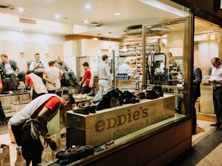 A busy shoeshine and repair store featuring multiple people getting their shoes shined by workers wearing red shirts. Shelves filled with shoes and repair equipment are visible, along with a storefront displaying 'Eddie’s Shoeshine & Repair'. The atmosphere is lively with a mix of customers seated and staff attending to them.