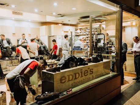 A busy shoeshine and repair store featuring multiple people getting their shoes shined by workers wearing red shirts. Shelves filled with shoes and repair equipment are visible, along with a storefront displaying 'Eddie’s Shoeshine & Repair'. The atmosphere is lively with a mix of customers seated and staff attending to them.