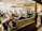 A busy shoeshine and repair store featuring multiple people getting their shoes shined by workers wearing red shirts. Shelves filled with shoes and repair equipment are visible, along with a storefront displaying 'Eddie’s Shoeshine & Repair'. The atmosphere is lively with a mix of customers seated and staff attending to them.