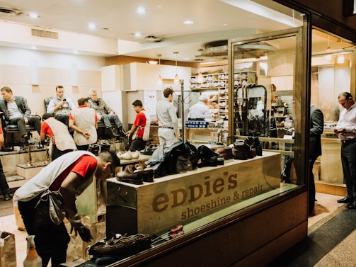 A busy shoeshine and repair store featuring multiple people getting their shoes shined by workers wearing red shirts. Shelves filled with shoes and repair equipment are visible, along with a storefront displaying 'Eddie’s Shoeshine & Repair'. The atmosphere is lively with a mix of customers seated and staff attending to them.