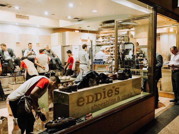 A busy shoeshine and repair store featuring multiple people getting their shoes shined by workers wearing red shirts. Shelves filled with shoes and repair equipment are visible, along with a storefront displaying 'Eddie’s Shoeshine & Repair'. The atmosphere is lively with a mix of customers seated and staff attending to them.