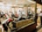 A busy shoeshine and repair store featuring multiple people getting their shoes shined by workers wearing red shirts. Shelves filled with shoes and repair equipment are visible, along with a storefront displaying 'Eddie’s Shoeshine & Repair'. The atmosphere is lively with a mix of customers seated and staff attending to them.