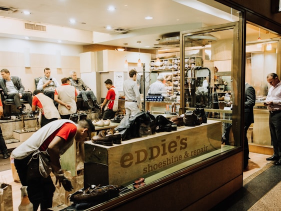 A busy shoeshine and repair store featuring multiple people getting their shoes shined by workers wearing red shirts. Shelves filled with shoes and repair equipment are visible, along with a storefront displaying 'Eddie’s Shoeshine & Repair'. The atmosphere is lively with a mix of customers seated and staff attending to them.