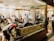 A busy shoeshine and repair store featuring multiple people getting their shoes shined by workers wearing red shirts. Shelves filled with shoes and repair equipment are visible, along with a storefront displaying 'Eddie’s Shoeshine & Repair'. The atmosphere is lively with a mix of customers seated and staff attending to them.