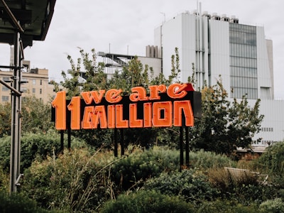A neon sign with the text '11 we are MILLION' stands amidst lush greenery, with modern urban buildings in the background.