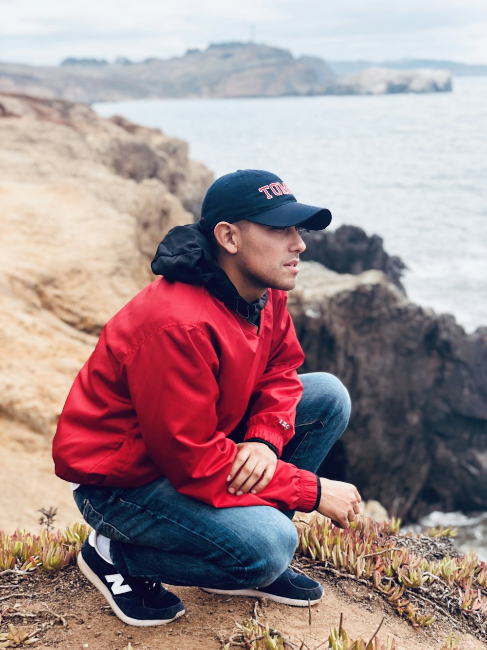 man wearing cap and jacket squatting on top of the cliff looking the overview