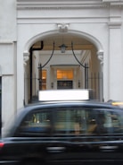 A black taxi is motion blur passing in front of a classical architectural archway. The archway features decorative molding and wrought iron elements. Inside the arch, there are glowing lights and a hallway leading further inside.