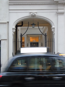 A black taxi is motion blur passing in front of a classical architectural archway. The archway features decorative molding and wrought iron elements. Inside the arch, there are glowing lights and a hallway leading further inside.