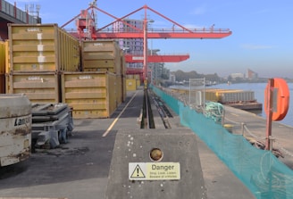 A shipping dock with stacked yellow containers labeled 'CORY' is adjacent to a body of water. A large red crane looms overhead, while a concrete barrier with a warning sign is in the foreground. Several industrial elements, such as machinery and cables, are present on the dock. Buildings can be seen in the distance, beyond the water.