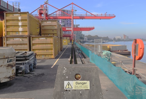 A shipping dock with stacked yellow containers labeled 'CORY' is adjacent to a body of water. A large red crane looms overhead, while a concrete barrier with a warning sign is in the foreground. Several industrial elements, such as machinery and cables, are present on the dock. Buildings can be seen in the distance, beyond the water.
