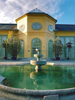 A symmetrical building with a yellow facade and large arched windows stands behind a circular stone fountain. The fountain is filled with water, reflecting the building and surrounding trees. The sky is partly cloudy, creating a bright, serene overall atmosphere.