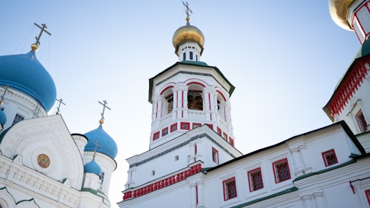 A scenic view of an ornate Orthodox church featuring vivid blue domes adorned with golden crosses. The central structure exhibits detailed white walls with red and green accents, and is capped with a golden dome. Architectural elements such as arches, windows, and decorative icons are visible, enhancing the church's intricate design.