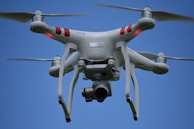 A white quadcopter drone is flying against a clear blue sky. It has four rotor blades, visible red markers on its arms, and a camera mounted underneath.