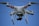 A white quadcopter drone is flying against a clear blue sky. It has four rotor blades, visible red markers on its arms, and a camera mounted underneath.