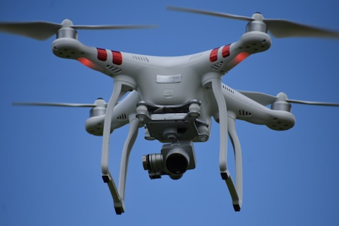 A white quadcopter drone is flying against a clear blue sky. It has four rotor blades, visible red markers on its arms, and a camera mounted underneath.