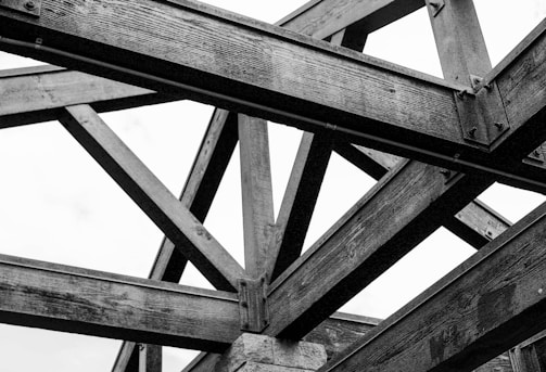 An intricate pattern of wooden beams creates a triangular lattice structure. The aged wood exhibits a rugged texture, and metal bolts secure the joints. The composition displays geometric symmetry and strong lines against a white background.