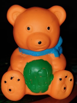 An orange teddy bear toy with black eyes and a small black nose sits against a dark background. The bear wears a blue scarf around its neck and features a green patch on its stomach with the words 'I love my teddy' inscribed on it.