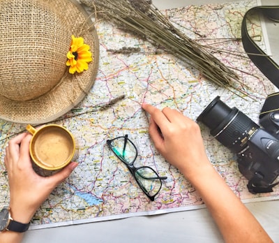 A colorful map is spread out with a straw hat decorated with a yellow flower, a mug of coffee, glasses, a camera, and a hand pointing to a location. Dried flowers are arranged at the top of the map.