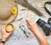 A colorful map is spread out with a straw hat decorated with a yellow flower, a mug of coffee, glasses, a camera, and a hand pointing to a location. Dried flowers are arranged at the top of the map.