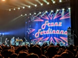 A large, excited crowd is gathered in front of a stage where a band is about to perform. The backdrop is colorful, featuring neon-style text that reads 'Franz Ferdinand' surrounded by star and arrow designs. The stage is brightly lit with spotlights shining overhead.