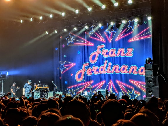 A large, excited crowd is gathered in front of a stage where a band is about to perform. The backdrop is colorful, featuring neon-style text that reads 'Franz Ferdinand' surrounded by star and arrow designs. The stage is brightly lit with spotlights shining overhead.