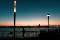 A smiling woman wearing headphones, jogging along a city waterfront at sunset.