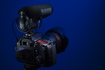 A DSLR camera equipped with a RODE microphone mounted on top is positioned against a solid blue background. The camera's display screen is visible, along with various control buttons and dials. The microphone is prominently branded with the RODE logo.
