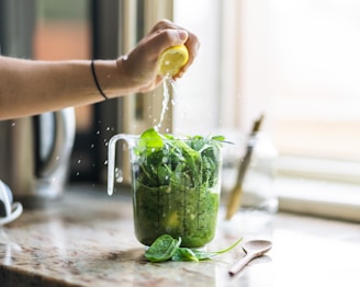 A hand squeezing a lemon over a measuring cup filled with fresh green spinach on a kitchen countertop. Lemon juice splashes over the leafy greens, and a wooden spoon rests nearby. The background features blurred kitchen elements.