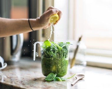 A hand squeezing a lemon over a measuring cup filled with fresh green spinach on a kitchen countertop. Lemon juice splashes over the leafy greens, and a wooden spoon rests nearby. The background features blurred kitchen elements.