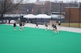 A group of lacrosse players in action on a green field with a goal net. They are wearing protective gear and actively engaged in practice or a game. The background features banners, a fence, and trees, suggesting an organized sports facility.