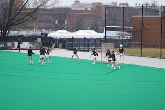 A group of lacrosse players in action on a green field with a goal net. They are wearing protective gear and actively engaged in practice or a game. The background features banners, a fence, and trees, suggesting an organized sports facility.