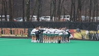 A group of athletes huddled together in a circle on a green sports field. They are wearing uniforms with green shorts and white tops, indicating a team sport environment. Lacrosse sticks are lined up against a fence in the background. Trees and parked cars are visible beyond the fence, suggesting an outdoor location.