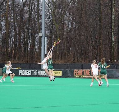 A group of lacrosse players on a green field, competing for the ball mid-air. The players are wearing green and white uniforms, with numbers visible on their jerseys. Bare trees and a banner with motivational text are in the background.