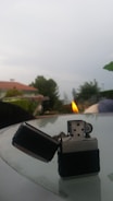 A metallic lighter with a black grip is lit on top of a glass table. The background features a blurred outdoor setting with trees, a house with a red roof, and a cloudy sky.